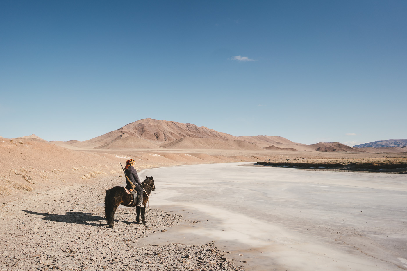 Mongolian farmer on horseback with gun over the shoulder looking over the frozen river and desert landscape of the Mongolian mountains. Captured by adventure photographer Stefan Haworth