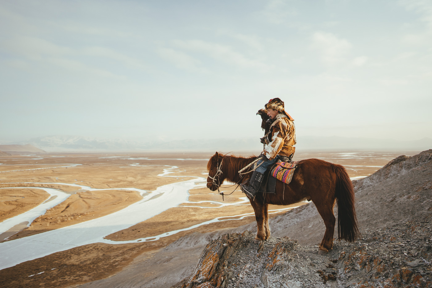 Kazakh eagle hunter on horseback looking over the frozen rivers in the valley with eagle and hand during sunset. Photographed by adventure photographer Stefan Haworth