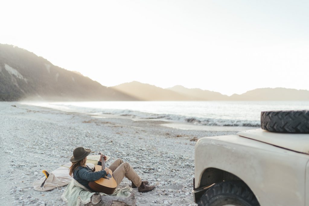 Photographer Stefan Haworth playing guitar at sunset beside the Series Land Rover in Jackson bay on the West coast of New Zealand.