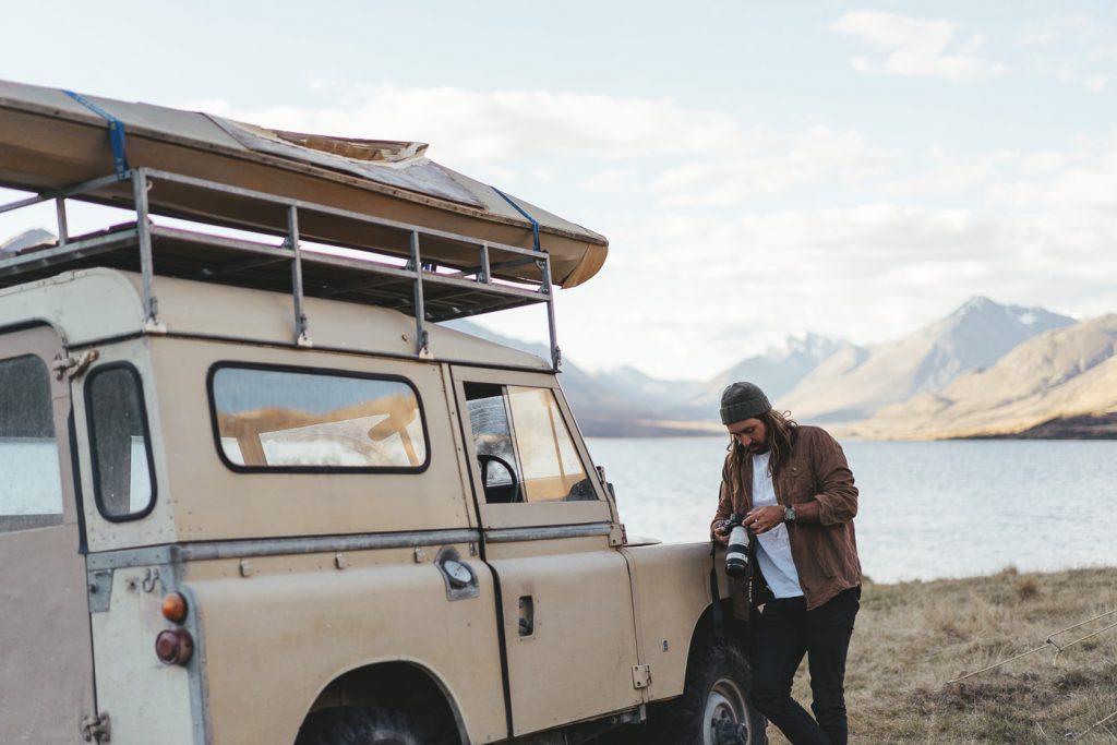 Photographer Stefan Haworth reviewing photos leaning against a Series Land Rover in remote lake of New Zealand's Southern Alps