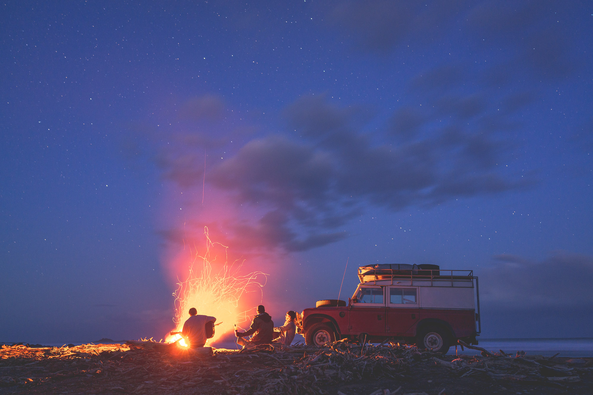 Friends sitting around a campfire playing guitar at sunset next to Land Rover defender in New Zealand